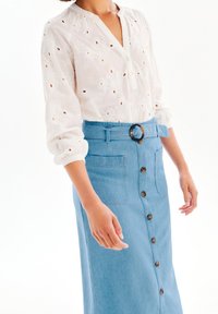 Vit broderad blus med v-ringning och lång ärm, i kombination med en ljusblå denimkjol med knäppning fram och ett brett bälte.