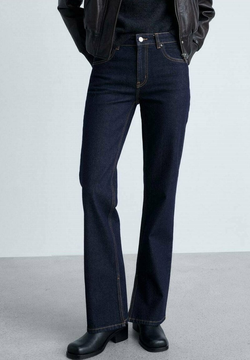 Personne portant un jean droit bleu foncé, des bottines noires, un pull noir et une veste en cuir noire, les mains dans les poches.