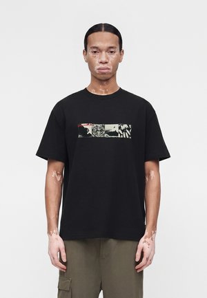 Άνδρας με λεύκη στέκεται με το πρόσωπο προς τα εμπρός, φοράει μαύρο γραφικό μπλουζάκι και ελαιδί παντελόνια cargo, μπροστά σε απλό φωτεινό φόντο.