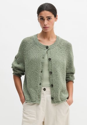 Frau trägt hellgrünen, strukturierten, knöpfbaren Cardigan über einem gerippten Tanktop mit beiger Hose, steht mit den Händen in den Taschen vor weißem Hintergrund.