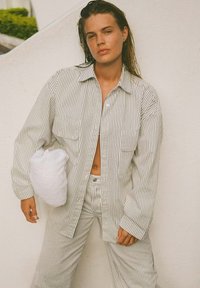 Gestreepte oversized blouse en broek in wit en lichtgrijs, met twee voorkeurszakken. Model houdt een witte fluffy kussentass accesoires vast.