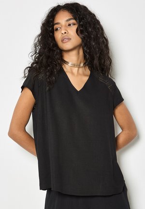 Vrouw met lang krullend haar, gekleed in een zwart V-hals shirt met korte mouwen en kanten details, met een gouden choker ketting, kijkt opzij.