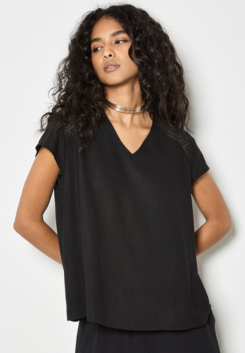 Femme aux longs cheveux bouclés portant un haut noir à col en V et manches courtes avec des détails en dentelle et un collier ras-du-cou en or, regardant sur le côté.