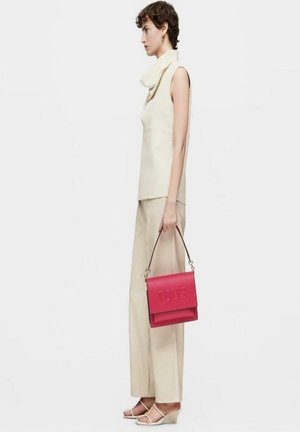 Bolso de piel rosa con el logo "TOUS" en relieve, sostenido por una persona con un atuendo en crema y sandalias de cuña blancas. Textura suave y diseño estructurado.