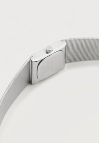 Κοντινή λήψη ασημένιου μεταλλικού ιμάντα ρολογιού και της πίσω πλευράς ορθογώνιας κάσας ρολογιού με χαραγμένο κείμενο και περιστρεφόμενο κουμπί.