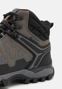 Braune Wildleder- und schwarze synthetische Wanderschuhe mit blauer Nähte, ausgestattet mit einem wasserdichten Etikett, robustem Schnürsystem und rutschfester Sohle.