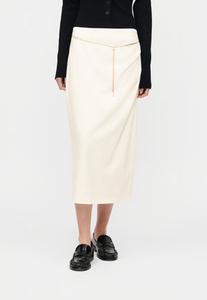 Γυναίκα που φοράει κρεμ μίντι φούστα με λεπτή χρυσή αλυσίδα στη μέση, μαύρη μπλούζα με μακριά μανίκια και μαύρα δερμάτινα μοκασίνια σε λευκό φόντο.