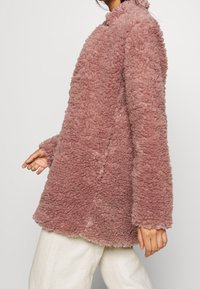 Tøff, oversized lyserød frakke lavet af blødt, tekstureret stof med høj krave og lange ærmer, der har et lige snit og minimal syning.