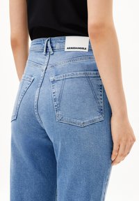 Los jeans de mezclilla azul claro tienen una cintura alta, dos bolsillos traseros y una etiqueta visible en la cinturilla con el nombre de la marca "ARMEDANGELS."