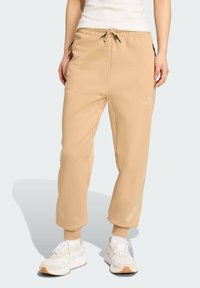 Béžové jogger nohavice z mäkkého materiálu s elastickým pásom a šnúrkou. Obsahujú bočné zipsové vrecká a nenápadné logo pri boku.