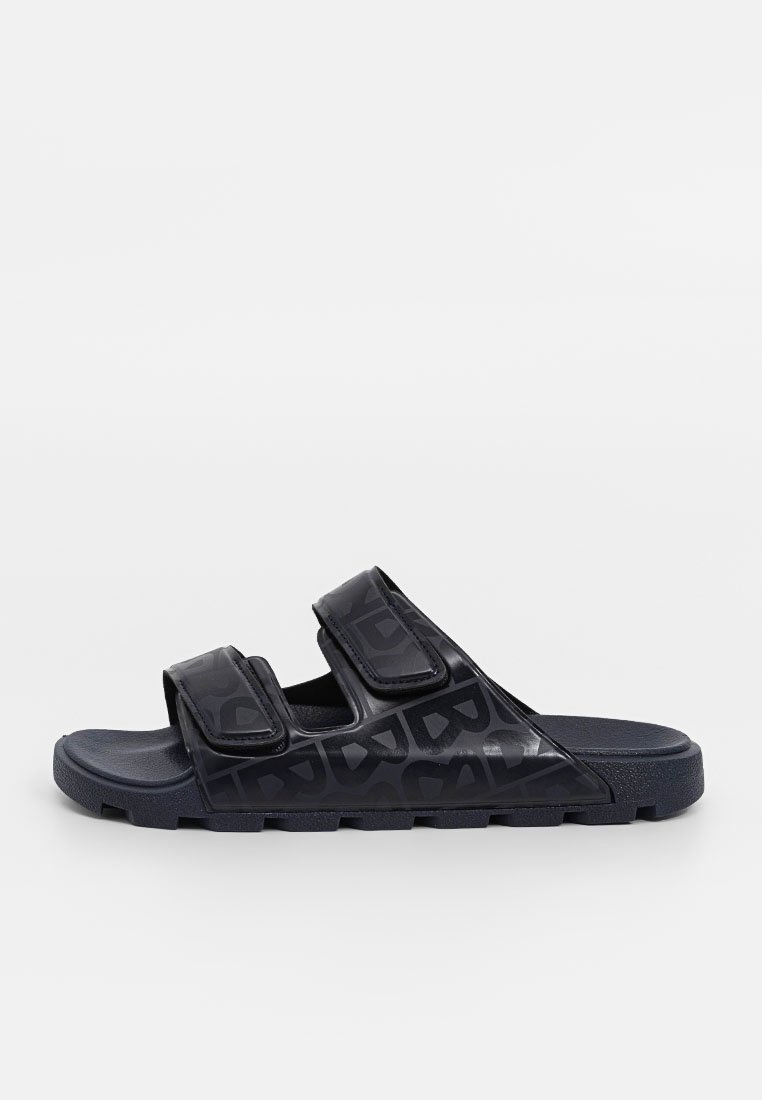 Schwarze Rutschen mit doppelten Klettverschlussriemen, strukturiertem Gummimaterial, flacher Sohle und geprägtem Logo-Detail auf dem Obermaterial.