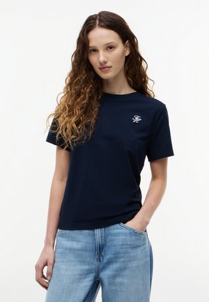 Νεαρή γυναίκα με μακριά σγουρά μαλλιά, φορούσα μπλε σκούρο μπλουζάκι με λευκό λογότυπο και ανοιχτόχρωμο μπλε τζιν, με το ένα χέρι στην τσέπη, ουδέτερο φόντο.