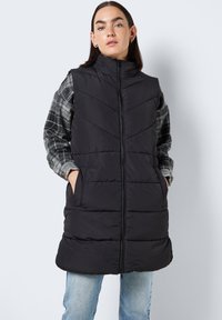 Svart polstret vest med høy krage, med kviltet chevrondesign, glidelås foran og to side lommer, lagd over en grå rutete skjorte.
