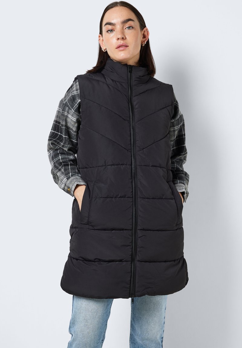 Svart polstret vest med høy krage, med kviltet chevrondesign, glidelås foran og to side lommer, lagd over en grå rutete skjorte.