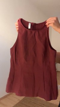 Due mani che tengono un top fitted senza maniche di colore bordeaux con scollatura alta contro uno sfondo neutro indoor.