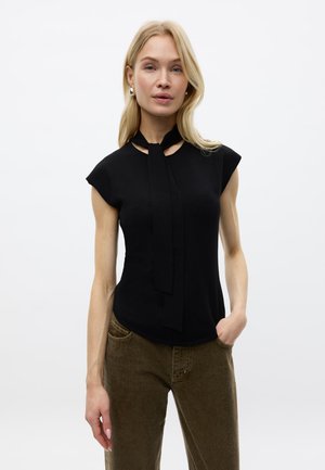 Vrouw met blond haar draagt een zwart mouwloos topje met stropdasdetail en een bruine broek, staande met één hand in de zak tegen een witte achtergrond.