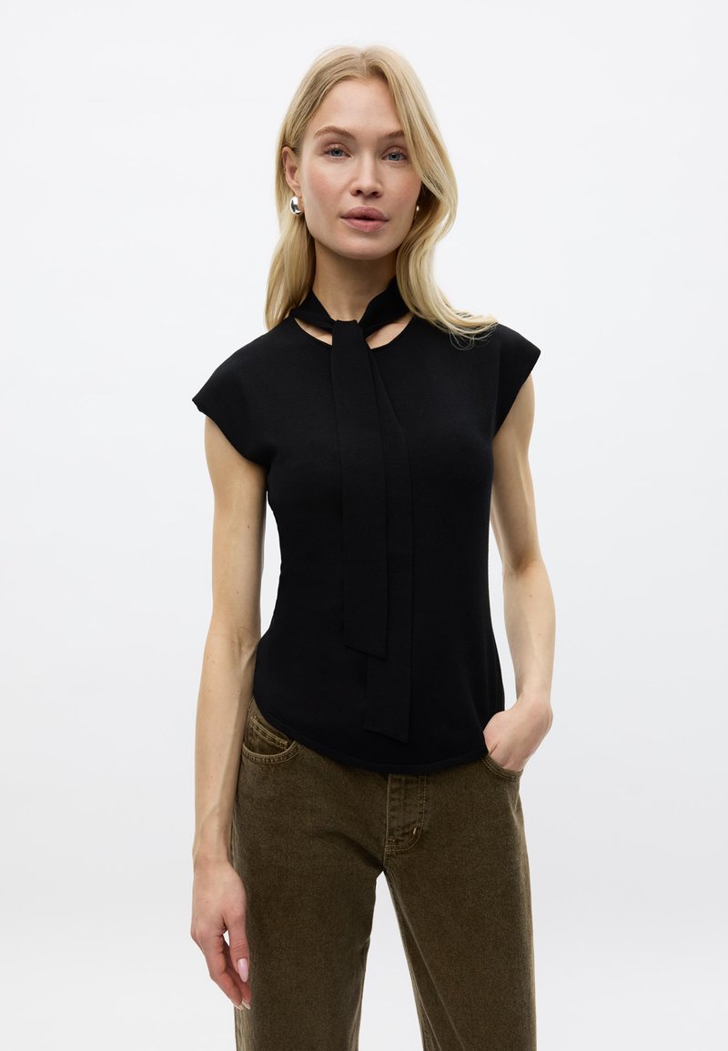 Femme aux cheveux blonds porte un top noir sans manches avec un détail cravate et un pantalon marron, debout avec une main dans la poche sur fond blanc.