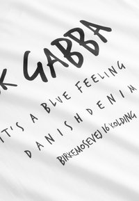 Bela bomba majica iz bombažne tkanine s krepko črnim besedilom. Besedilo vključuje »Gabba«, »It's a blue feeling« in naslov, v različnih velikostih in slogih pisave.