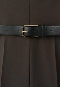 Cintura nera in pelle con fibbia dorata lucida, cinturino testurizzato, su sfondo di tessuto marrone con pieghe.