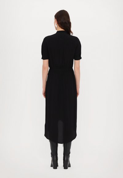 Rochie neagră, până la genunchi, cu mâneci scurte, talie strânsă și slit pe spate. Textură netedă cu o siluetă elegantă, purtată cu cizme până la genunchi.