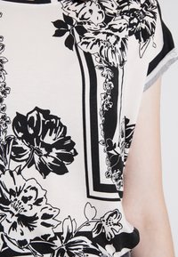 Svartvit blommig topp med korta ärmar. Den har en rund halsringning och en texturerad tyg med ett rektangulärt kantdesign.