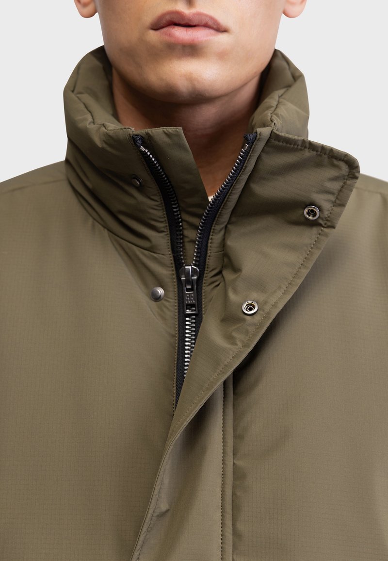 Giacca verde oliva con colletto alto, dotata di chiusura con zip, bottoni a scatto e tessuto testurizzato. Dettagli di cucitura limitati e visibili.