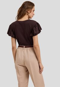 Brown ruffled short-sleeve shirt paired with beige high-waisted pants with an elastic waistband, seen from a slightly turned perspective.