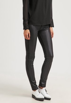 Pantalones negros de cuero ajustados combinados con una blusa negra transparente y zapatos de plataforma blancos con cremallera sobre un fondo liso.