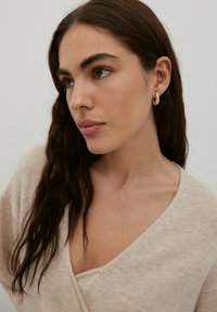 Pull en tricot beige avec un décolleté en V profond. Le modèle porte des boucles d'oreilles en or rondes. Les cheveux sont longs et ondulés, sur un fond clair.