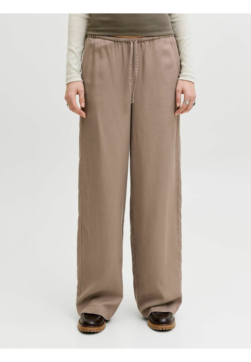 Personne portant un pantalon ample à cordon de serrage couleur taupe et des chaussures marron foncé, debout contre un fond clair uni.