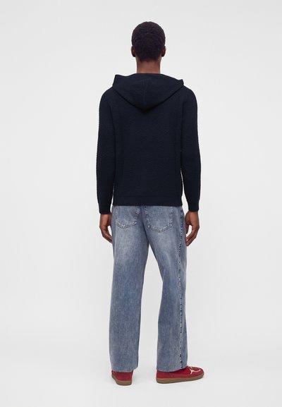 Homme portant un sweat à capuche noir, un jean bleu clair et des baskets rouges, debout de dos sur un fond clair uni.