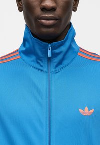 Μπλε ζακέτα με φερμουάρ και ψηλό γιακά, με πορτοκαλί τριπλές ρίγες στους ώμους και κεντημένο λογότυπο στο στήθος. Λείο ύφασμα.