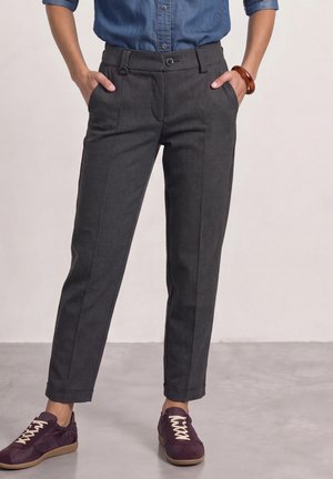 Person trägt dunkelgraue karierte Hose, blaues Hemd mit Knöpfen, lila Turnschuhe und ein braunes Armband mit den Händen in den Taschen.