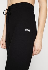 Pantalones de chándal de algodón negro con cintura con cordón, que presentan un bordado plateado de "DKNY" en la cadera izquierda. Textura suave, ajuste relajado.