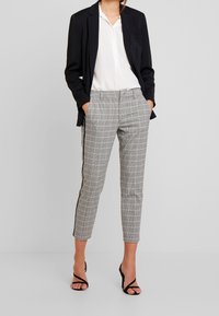 Pantalon à carreaux gris et blancs, coupe slim, poches latérales, bandes noires sur les côtés et ourlet retroussé, associé à un blazer noir.