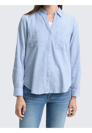 Frau trägt hellblaues, strukturiertes Langarmhemd mit Knöpfen und zwei Brusttaschen, kombiniert mit mittelblauen Jeans.