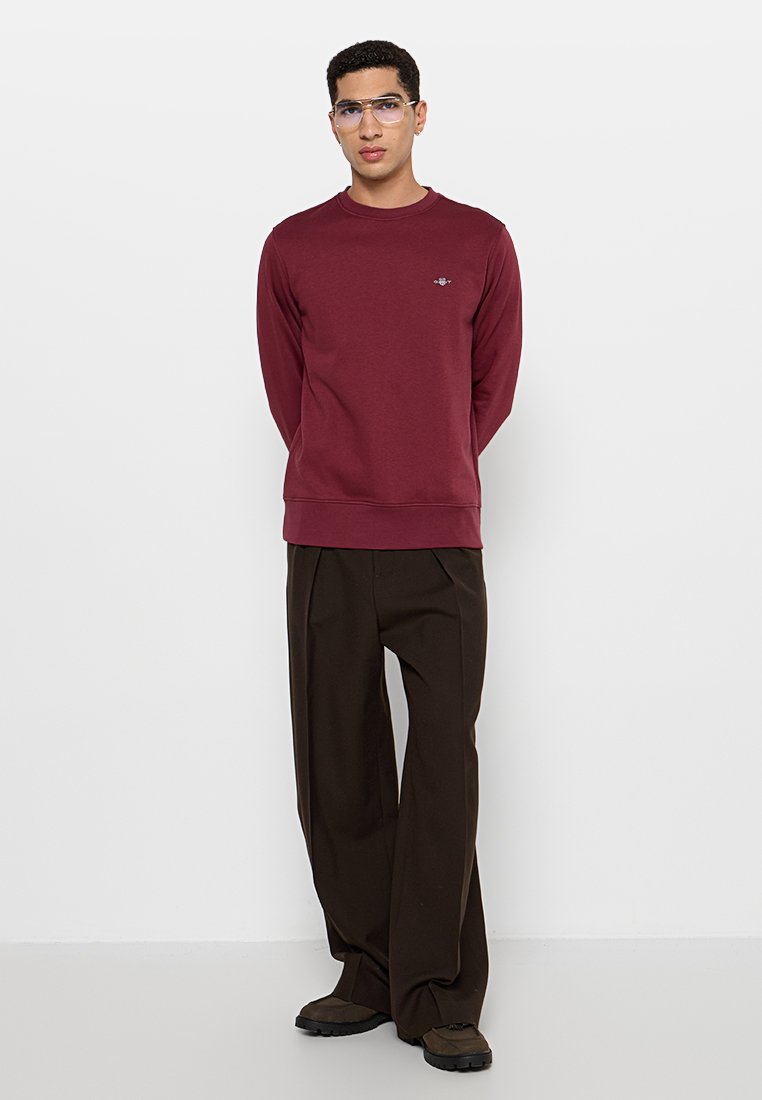 Sudadera burdeos con cuello redondo, mangas largas y puños acanalados, combinada con pantalones marrones de corte holgado y calzado negro.