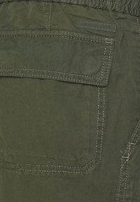 Olivgrüne Stoffshorts mit einer Gesäßtasche und einem rechteckigen Patch, betont durch weiße Nähte und einem dezenten Logolabel.