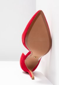 Chaussure à talon haut en suede rouge avec un bout pointu, un talon stiletto fin, et une semelle en cuir beige, présentée sur un fond blanc.