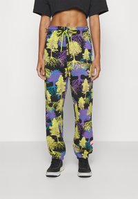 Person wearing black sweatpants with vibrant yellow and purple tree and root patterns, paired with black sneakers and a cropped black top.