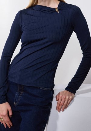 Vrouw draagt een aansluitende marineblauwe geribbelde longsleeve met een gouden sluiting bij de hals, gecombineerd met donkerblauwe spijkerbroek.