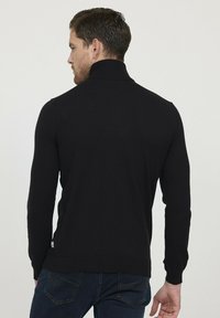 Pull en tricot noir avec un col montant, des manches longues et des poignets côtelés. Notable pour sa texture lisse et sa coupe ajustée.