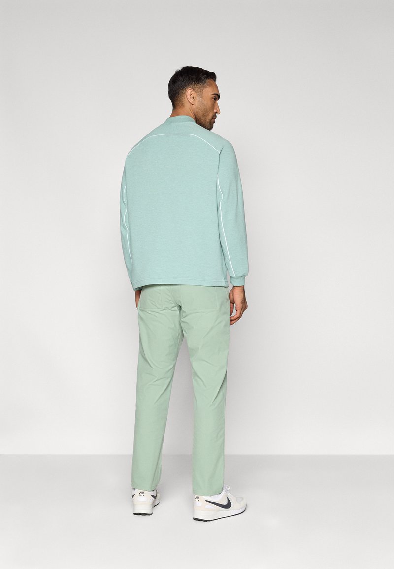 Lichtgroene trui met witte accenten, gecombineerd met bijpassende lichtgroene broek. Model draagt witte sneakers, gezien van achteren.