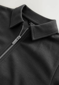Must vähem atrasiivse tekstuuriga must zip-up sviiter, millel on krae ja metallist lukukinnitus. Silt ütleb "NEXT". Sile pind ribadega detailidega.