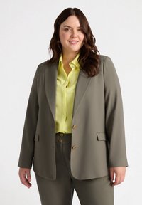 Sacou verde Kaley cu două butoane aurii, asortat cu o cămașă galbenă strălucitoare, cu o textură fină și croi ajustat.
