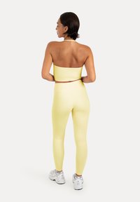 Femme aux cheveux courts et foncés, de dos, portant un haut bandeau jaune pâle, des leggings assortis, des baskets blanches et des chaussettes blanches sur fond blanc.