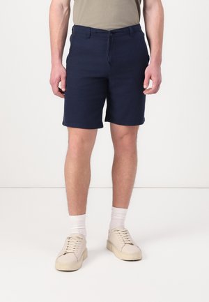 Uomo che indossa pantaloncini blu navy, t-shirt beige, calzini bianchi e scarpe da ginnastica beige, in piedi su un pavimento bianco contro un muro bianco.