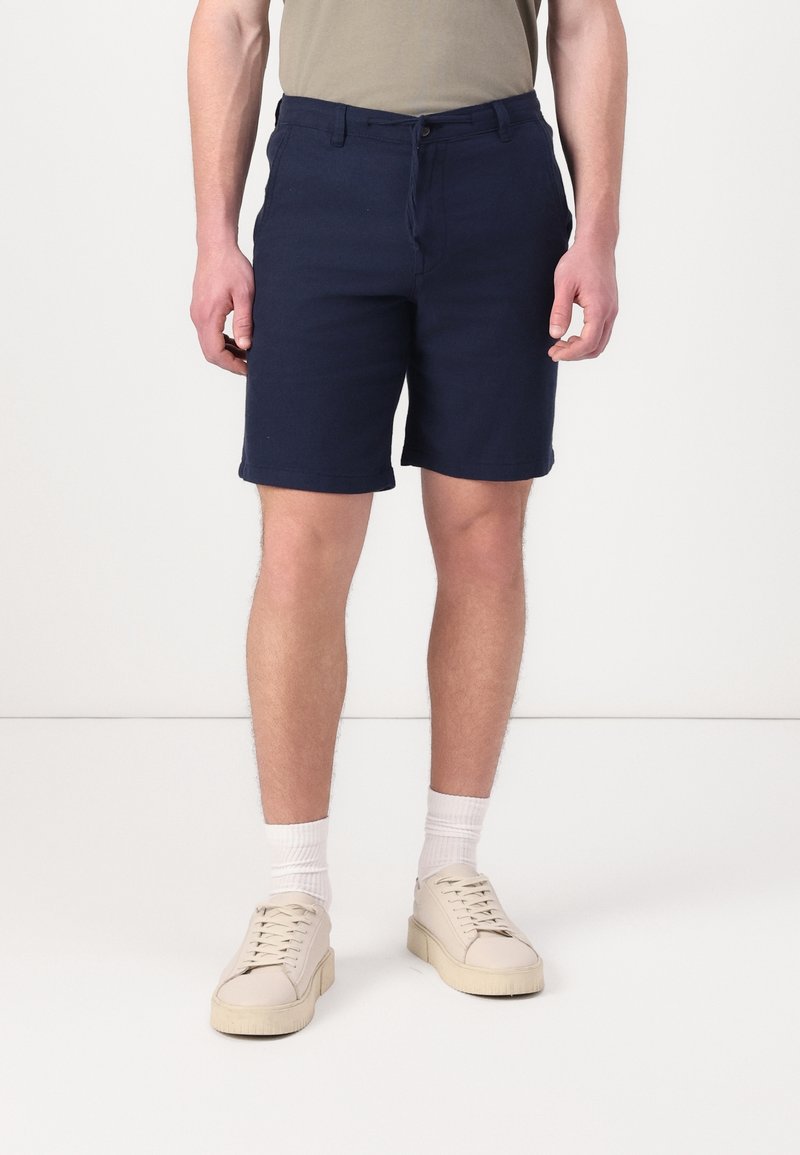 Homme portant un short bleu marine, un t-shirt beige, des chaussettes blanches et des baskets beiges, debout sur un sol blanc contre un mur blanc.