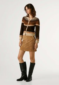 Cardigan lavorato a maglia marrone con motivo a zigzag, dettagli beige e bottoni. Abbinato a una gonna in corduroy color nocciola e stivali neri al ginocchio.
