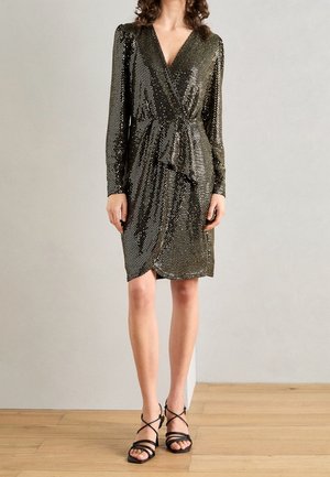 Kobieta w czarno-srebrnej metalicznej sukience kopertowej z długim rękawem, zestawiona z czarnymi sandałami na obcasie, stojąca na drewnianej podłodze.